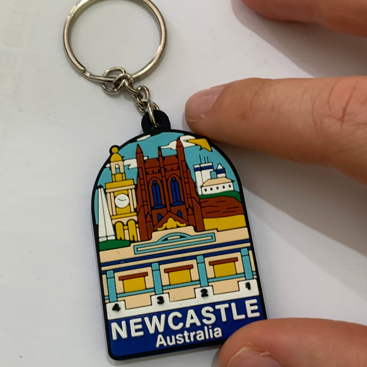 A Newcastle Australia souvenir keychain held by fingers against a plain white background. The keychain features a colorful illustration of Newcastle landmarks and the text 'NEWCASTLE Australia' on a blue base, attached to a silver metal key ring.